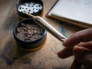 How to Roll the Perfect Joint in 5 Easy Steps 3 Hand holding rolled marijuana joint with grinder and cannabis herb on wooden table for recreational use